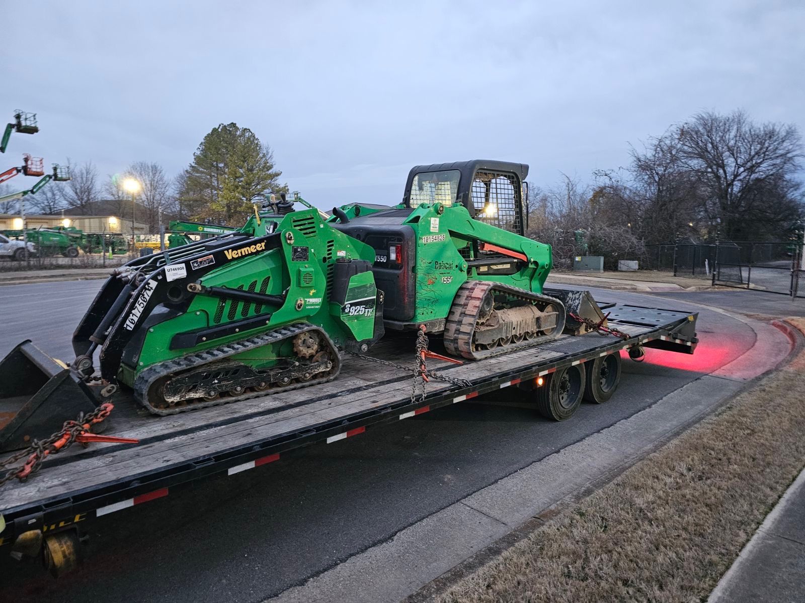 Two compact track loaders chained and strapped to a FlatbedLTL flatbed at dusk